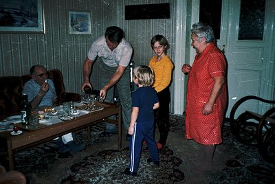 Family gathering in a rustic wooden interior, likely mid-20th century. A man in a white shirt and cap serves a young boy at a...