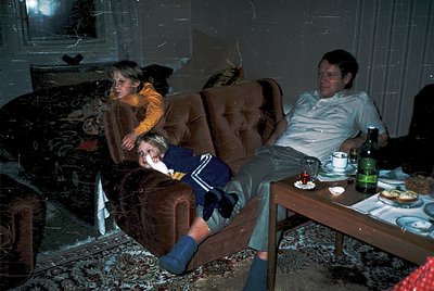 Vintage indoor scene featuring three individuals in a dimly lit living room, likely from the 1970s–1980s. Two men and a child...