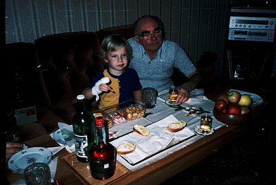 Vintage indoor family portrait from the 1970s–80s, featuring a man and child seated at a coffee table laden with food, wine b...