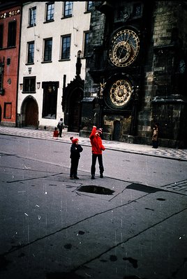Medieval astronomical clock mounted on Gothic-style building façade, featuring Roman numerals and zodiac imagery. Two figures...