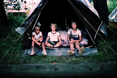 Three children pose inside a vintage canvas tent, seated on a rolled sleeping mat. The boy on the right wears a white T-shirt...