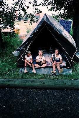 Three children sit inside a vintage canvas tent on a paved surface, surrounded by greenery. The tent’s patterned fabric and r...