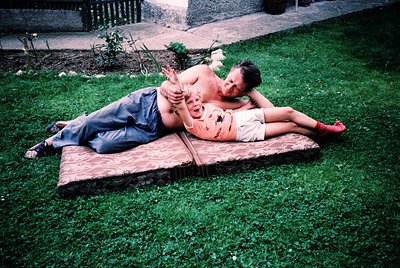 Vintage 1970s backyard scene: man in denim shorts reclines on a wooden bench, woman in floral dress and red shoes lies beside...