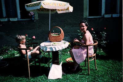 Mid-20th century garden scene with two adults and a child seated under a patterned umbrella. Woman in sleeveless dress and ma...