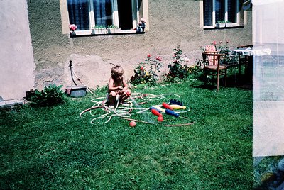 Vintage childhood scene: child playing with garden hose and inflatable toys on lush green lawn. Grey stucco house with white-...