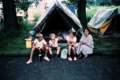 Family of four poses outside a canvas tent in a wooded campsite, mid-1970s–1980s. Three children and an adult woman sit on a ...