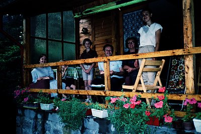 Five adults pose on a wooden balcony adorned with potted flowers and patterned curtains, likely a 1970s seaside resort. Rusti...