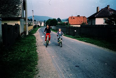 Two children riding bikes on a rural road, likely Eastern Europe, mid-20th century. The boy in red leads, followed by a girl ...