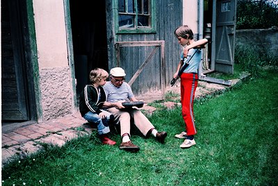 Vintage photo of three individuals in a rural courtyard, likely Eastern Europe, 1970s–80s. Elderly man in striped shirt and c...