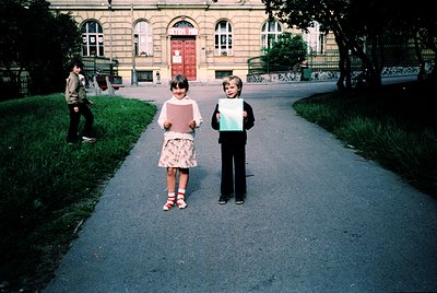 Two children pose holding vintage photo albums in a European urban park, 1970s. Girl in floral dress, boy in dark trousers. B...