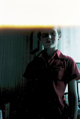 Portrait of a young boy in a maroon short-sleeve shirt with a collar, standing indoors near a window with visible condensatio...