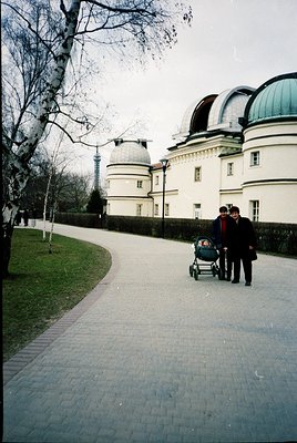 Mid-20th century courtyard scene featuring a white neoclassical building with domed observatory structures. Two adults push a...