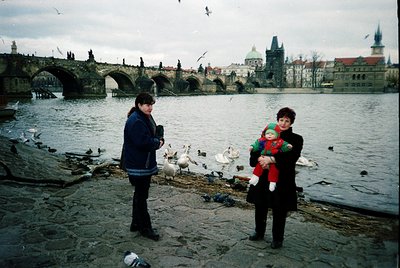Two women stand by the Vltava River in Prague, holding a child. The woman on the left wears a blue jacket, while the woman on...