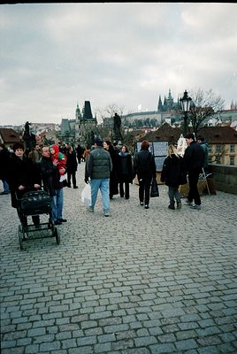 Cobblestone bridge bustling with pedestrians, likely Prague’s Charles Bridge, with historic Gothic architecture in background...