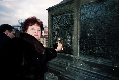 A woman in winter attire places a flower on a detailed bronze memorial plaque with engraved text and relief sculptures, likel...