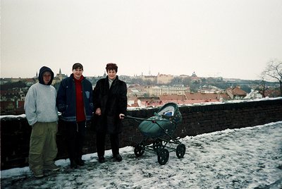 Three individuals pose on a snowy rooftop overlooking a historic European cityscape, likely Prague. The man on the left wears...