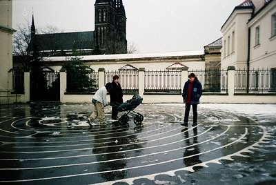 Three individuals in winter attire navigate a wet courtyard with circular pavement patterns. A historic church with a tall sp...