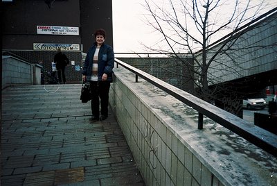 Woman in 1980s-style jacket and scarf stands on a concrete pedestrian bridge beside a "Fotocentrum" sign. Overcast urban sett...