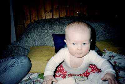 Vintage candid of an infant in a cozy indoor setting, seated on a patterned blanket with wooden paneling walls. The child wea...