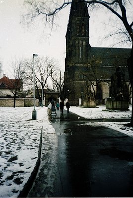 Historic European church tower with Gothic Revival details, flanked by bare winter trees and a statue. Snow-covered pavement ...