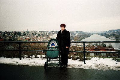 A woman in winter attire stands beside a vintage green stroller on a rooftop terrace overlooking a snow-covered urban landsca...