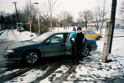 A woman in winter attire holds a child by a 1990s-era sedan in a snowy urban setting. Snow covers roads, parked cars, and sid...