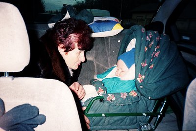 A woman in a vintage sweater cradles an infant in a stroller inside a car, surrounded by luggage and blankets. The scene sugg...
