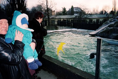 A family observes a yellow toy duck floating in a wave pool, likely at an indoor water park or aquarium. The adults wear wint...