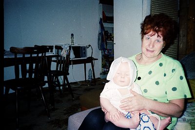 A woman in a polka-dot shirt cradles an infant in a dimly lit, utilitarian room. Wooden chairs and a cluttered shelf with bot...