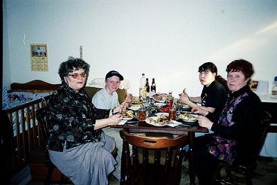 Four adults seated around a wooden table in a modest indoor setting, likely a 1990s-era home. The woman on the left wears pat...