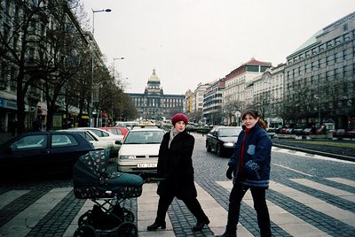Two women in winter attire cross a cobblestone pedestrian zone in a European city square, 1990s. The woman on the left wears ...