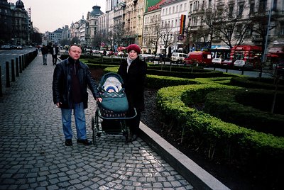 Couple strolling with a baby stroller on a cobblestone promenade in a European city, likely Prague. Classic 1990s-era clothin...
