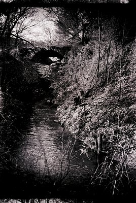 Vintage black-and-white photo of a rustic stone bridge spanning a shallow stream in a wooded area. The bridge’s archway frame...