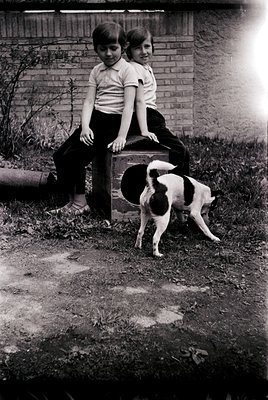 Two boys in 1970s-style polo shirts and pants pose outdoors beside a brick wall, leaning on a vintage suitcase. A black-and-w...