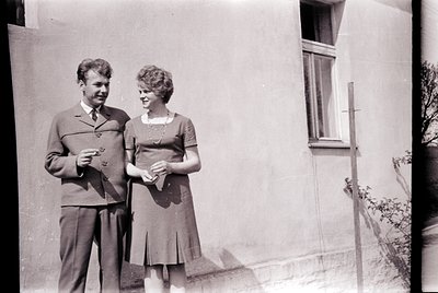 Couple posing outdoors in 1950s-60s attire, man in cardigan with tie, woman in knee-length dress with collar. Plain residenti...
