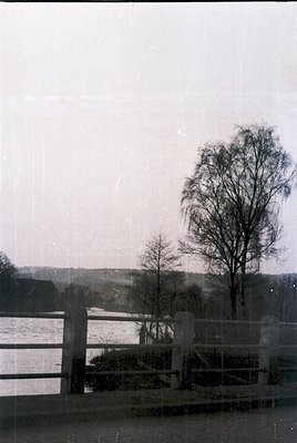 Vintage black-and-white landscape featuring a weathered wooden fence along a serene lake, framed by leafless trees and rollin...