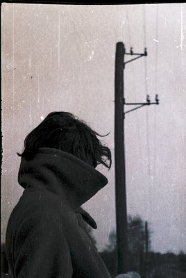 Silhouette of a person in a heavy winter coat and hat, back to camera, standing near a utility pole with crossbars. Blurred u...