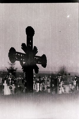 Ornate Orthodox-style cross silhouette against a cemetery backdrop, likely Eastern European. Intricate floral motifs on cross...