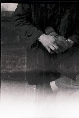 Close-up of a person’s legs and lower torso seated on a textured, possibly metal bench. The individual wears dark trousers, a...