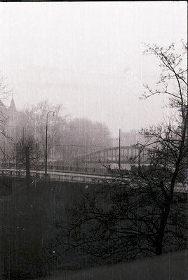 Fog-obscured urban scene featuring a bridge with vintage streetlamps and skeletal winter trees. Distant industrial buildings ...
