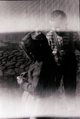 Vintage black-and-white photo of two children in outdoor setting, likely mid-20th century. One child wears a plaid shirt and ...