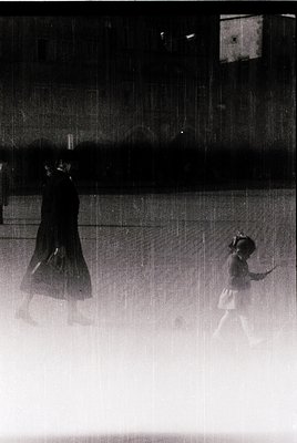 Blurred monochrome street scene capturing two figures in heavy rain. A woman in a long coat and hat walks with an umbrella, w...