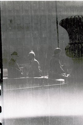 Reflection of three seated figures in a rain-soaked courtyard, viewed through a glass window. Distinctive classical column ba...