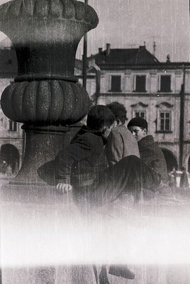 Three boys in 1950s-60s attire sit on ornate stone fountain base in urban plaza, rain obscuring background. Classic European ...