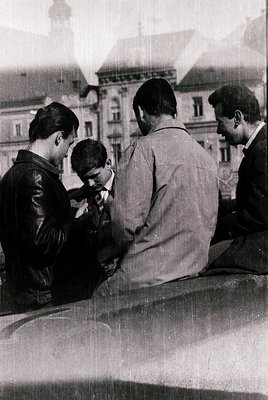 Three men in mid-20th-century attire (leather jacket, sweater, suit) huddle on a stone ledge, likely a balcony or terrace, in...