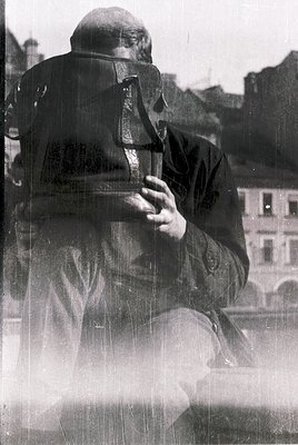 A man in mid-20th-century military gear—helmet, coat, and gas mask pouch—sits on a ledge overlooking an urban background. The...