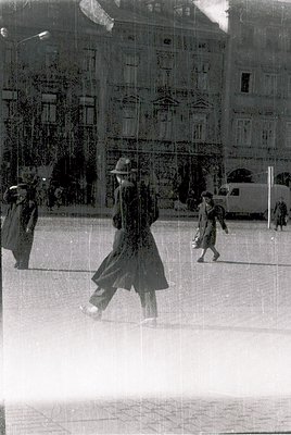Vintage monochrome street scene reflecting through a rain-smeared window. Central figure in a long coat and hat strides forwa...