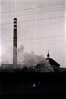 Industrial smokestack emitting haze beside a small church with a spire, framed by barbed wire. Mid-20th century urban/industr...