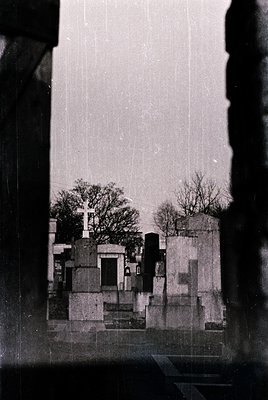 Mid-20th century cemetery view through a window frame, showcasing concrete mausoleums and leafless trees. Architectural Bruta...