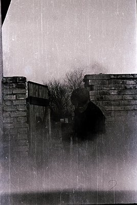 Vintage black-and-white photo of a lone figure standing behind a brick wall in heavy rain. The person wears a dark coat and h...
